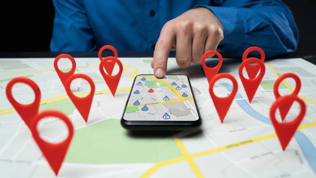 A person tapping a smartphone screen displaying a digital map, surrounded by large red location map pin markers set on a paper street map.