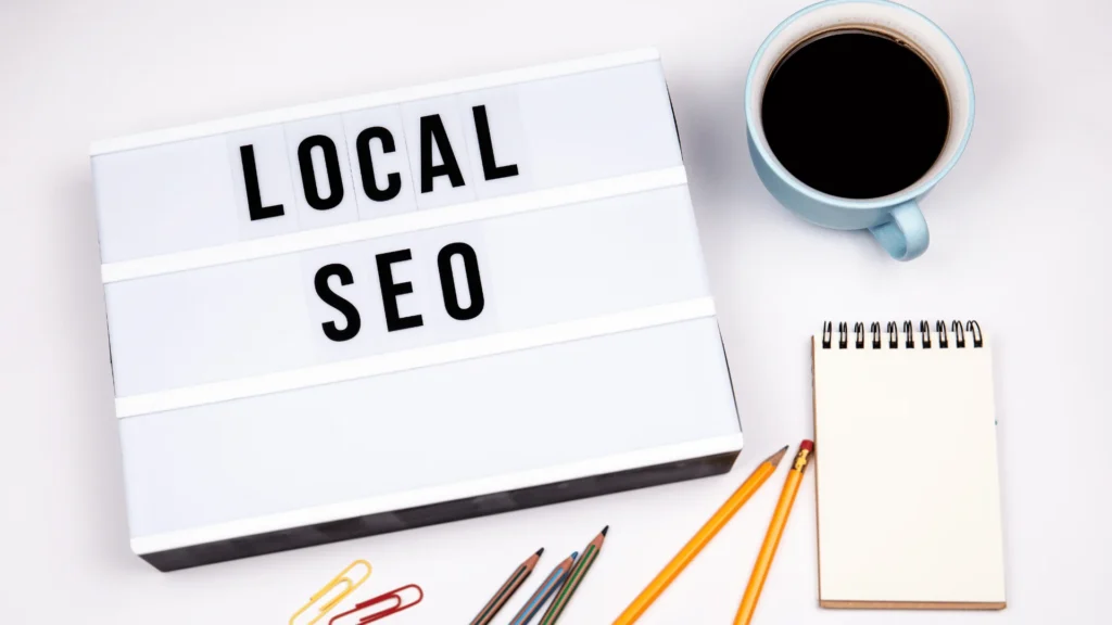 A flat-lay desk setup featuring a light-box sign reading "LOCAL SEO" beside a cup of black coffee, pencils, paper clips, and a blank spiral notepad.